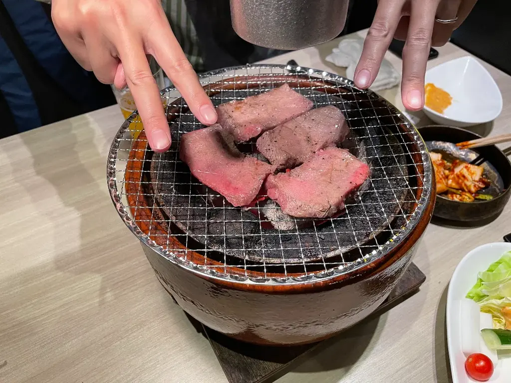 七輪の上に肉が四枚 ダブルピースしてる指が写り込んでいる 片手の血色が異常に悪く写っている