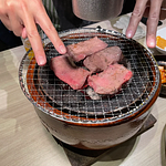 七輪の上に肉が四枚 ダブルピースしてる指が写り込んでいる 片手の血色が異常に悪く写っている